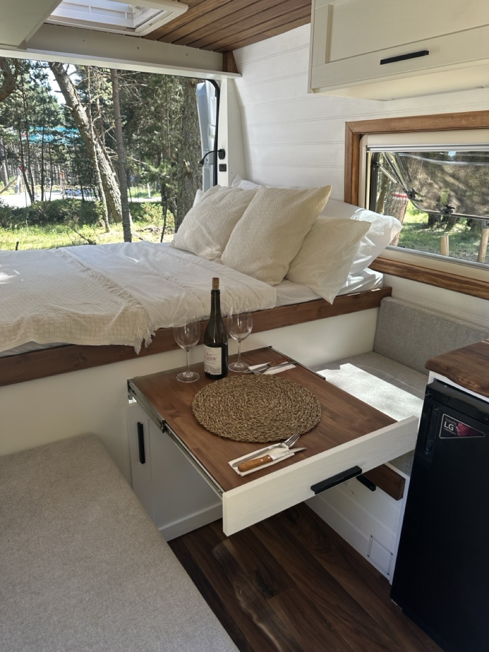 Interior de la camper FUGA con la cama preparada y una mesa con vino, junto a una ventana con vistas al bosque